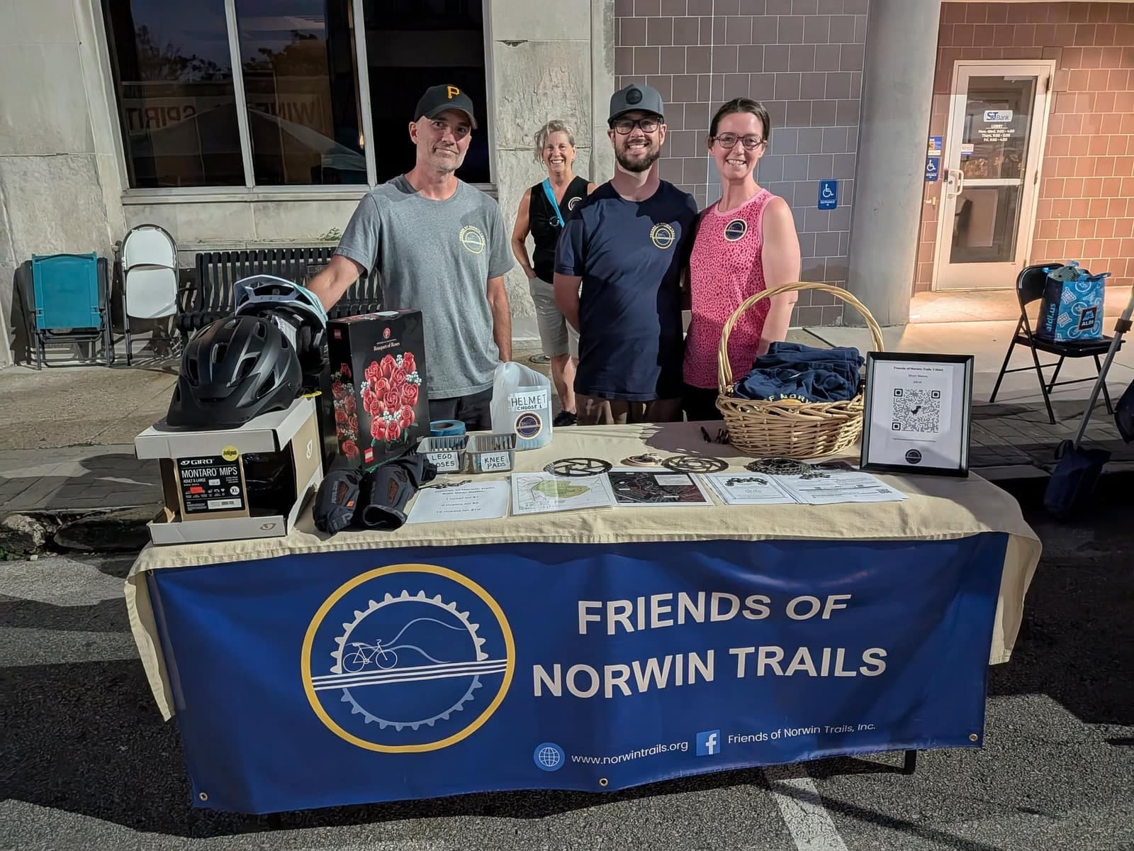 Volunteers behind a Friends of Norwin Trails banner at a fundraising table with raffle prizes, helmets, and t-shirts